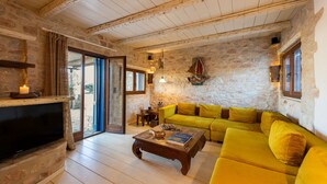 A cozy living room with a stone wall, a yellow sofa, a fireplace, and a television. - Blue Greens Villas in Krios, Paros Island (Paros)