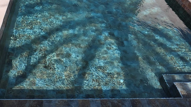 Ein rechteckiger, klarer Schwimmteich mit Glasboden, umgeben von einer gefliesten Terrasse.