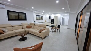 Ein modernes Wohnzimmer mit einem beigefarbenen Ecksofa, einem kleinen runden Couchtisch und einem braunen Sessel. Im Hintergrund ist eine Küche mit weißen Schränken sowie ein Essbereich mit Tisch und Stühlen zu sehen.
