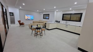 Ein modernes Wohnzimmer mit einer Couch, einem Esstisch mit Stühlen, einem an der Wand montierten Fernseher und einer Küchenzeile mit Schränken und einem Fenster.