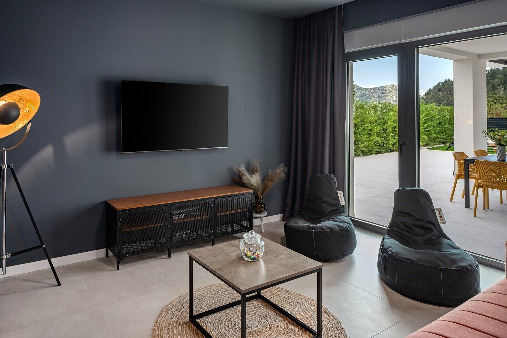 A modern living room with a flat-screen TV, a wooden bench, a coffee table, and a view of an outdoor patio with chairs and a table.