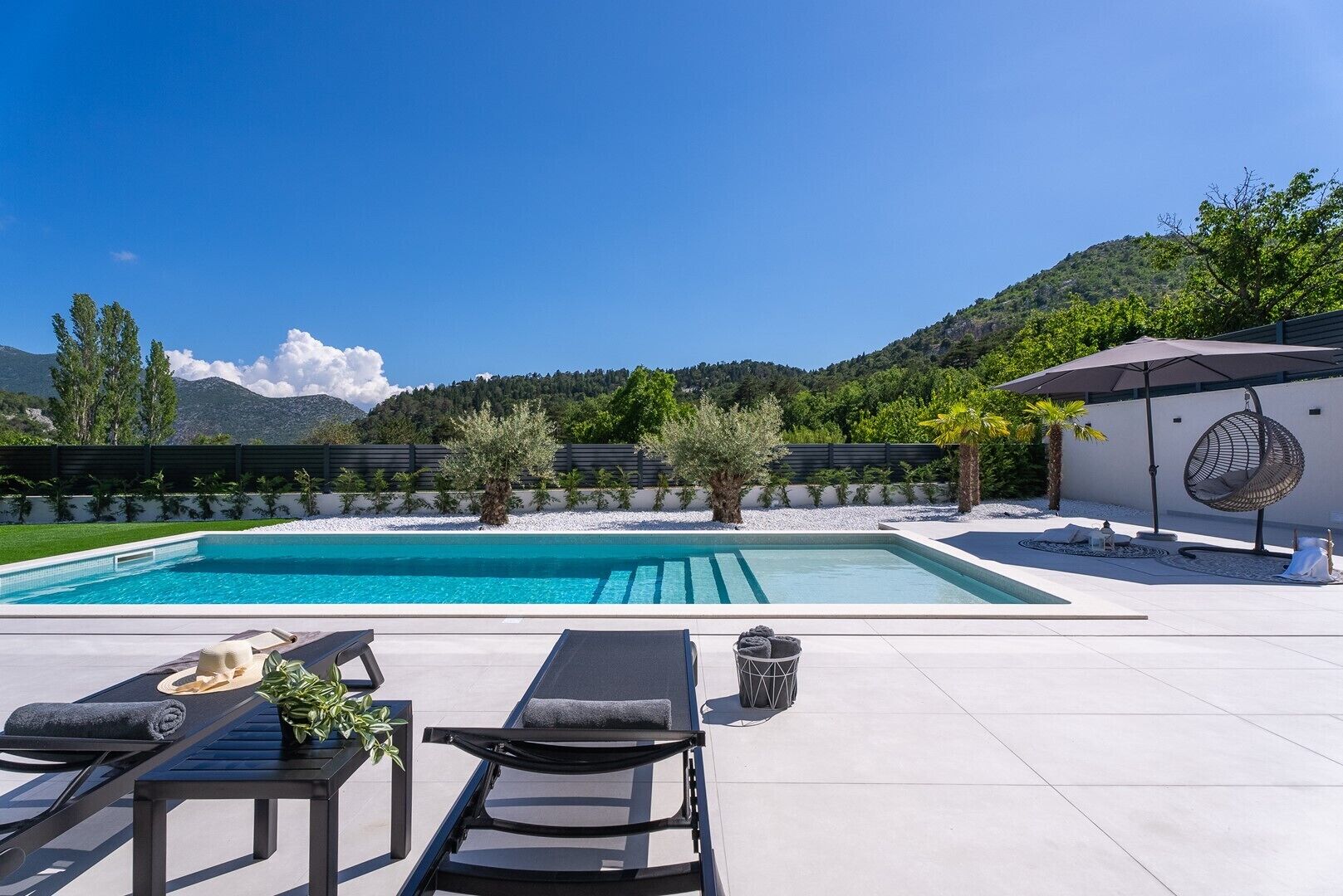 A swimming pool with lounge chairs and a hanging chair, surrounded by a landscaped garden and a view of mountains.