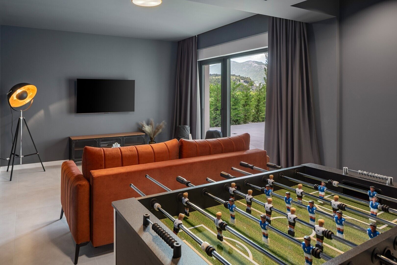 Una sala de estar moderna con mesa de fútbol, sofá, televisor y un ventanal con cortinas.