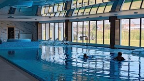 An indoor swimming pool with clear water, a few people swimming, and a view of the outdoors through large windows.