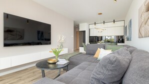 A modern living room with a large flat-screen TV, a sectional sofa, a small round table with a vase of flowers, and a kitchen area in the background.