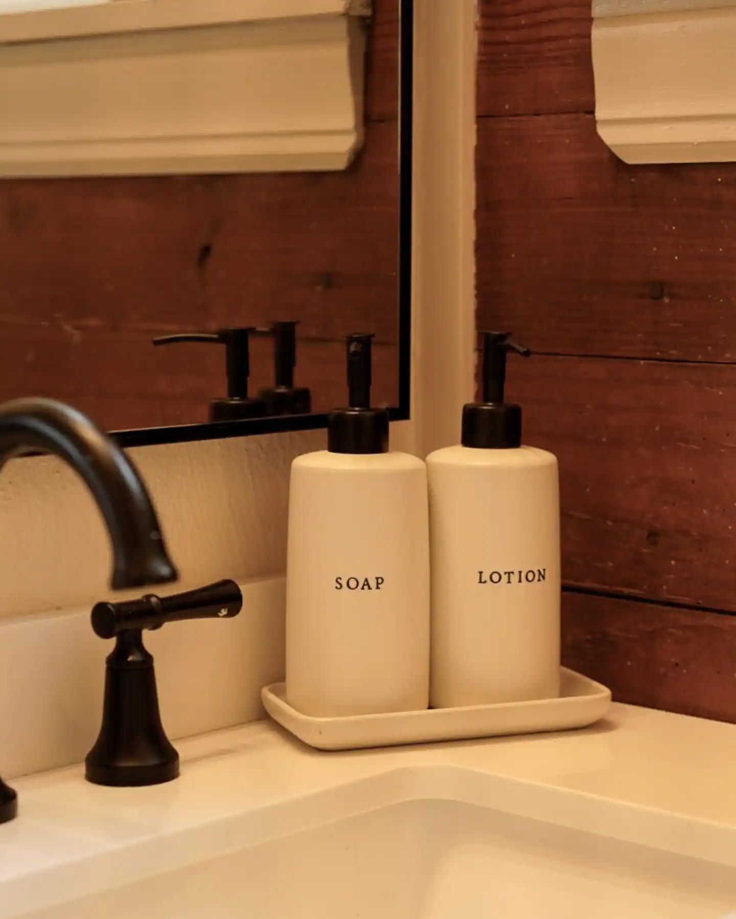 Un lavamanos con un grifo moderno, dos botellas beiges con los rótulos 'SOAP' y 'LOTION' sobre una bandeja, y una pared de madera al fondo.
