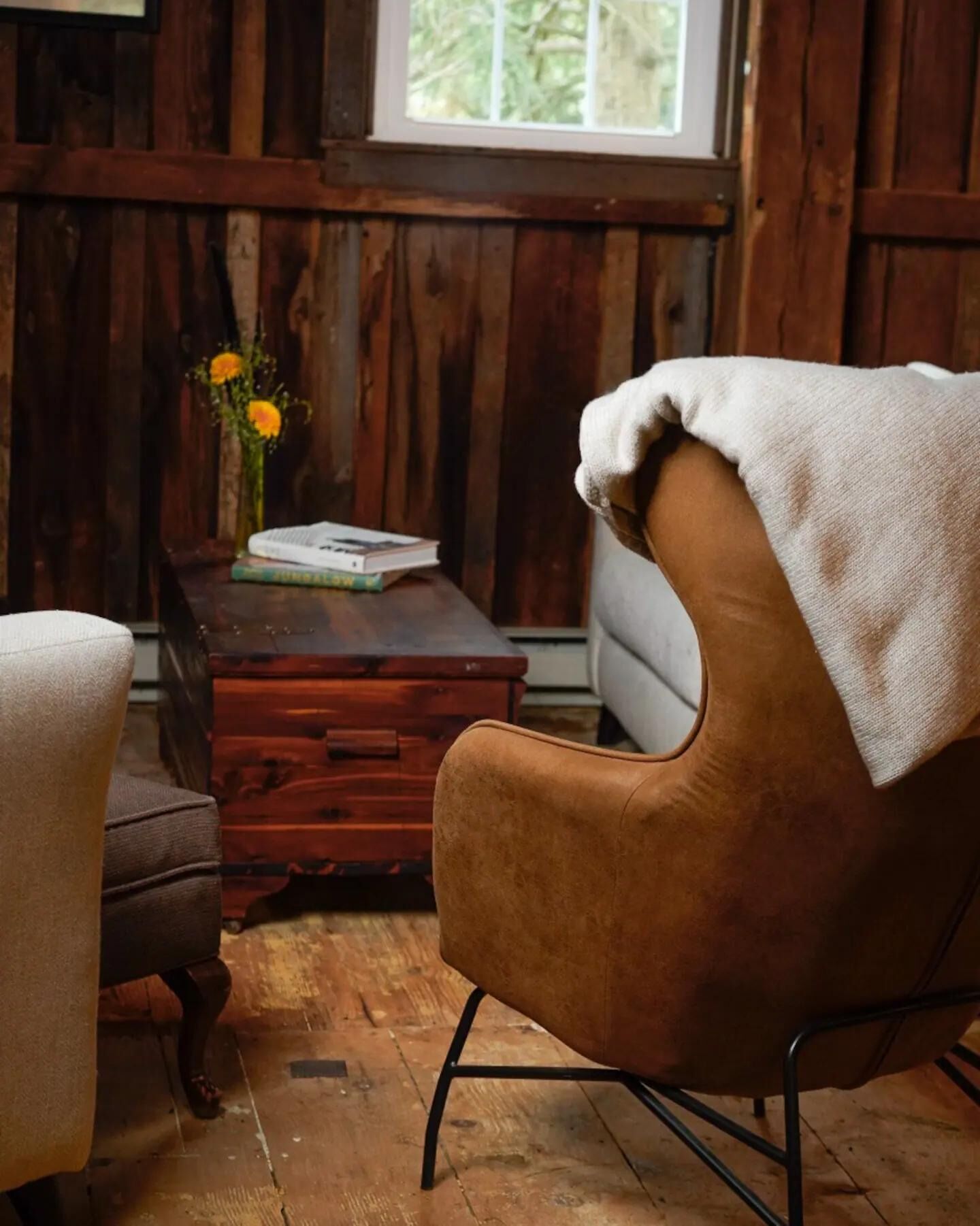 A rustic room with a brown leather chair, a wooden desk with a book and a vase of flowers, and a window with a view of trees.