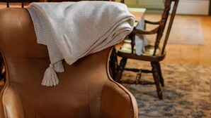 Una silla de cuero marrón con una manta blanca y flecos, una mesa de comedor de madera con sillas y una cocina al fondo.