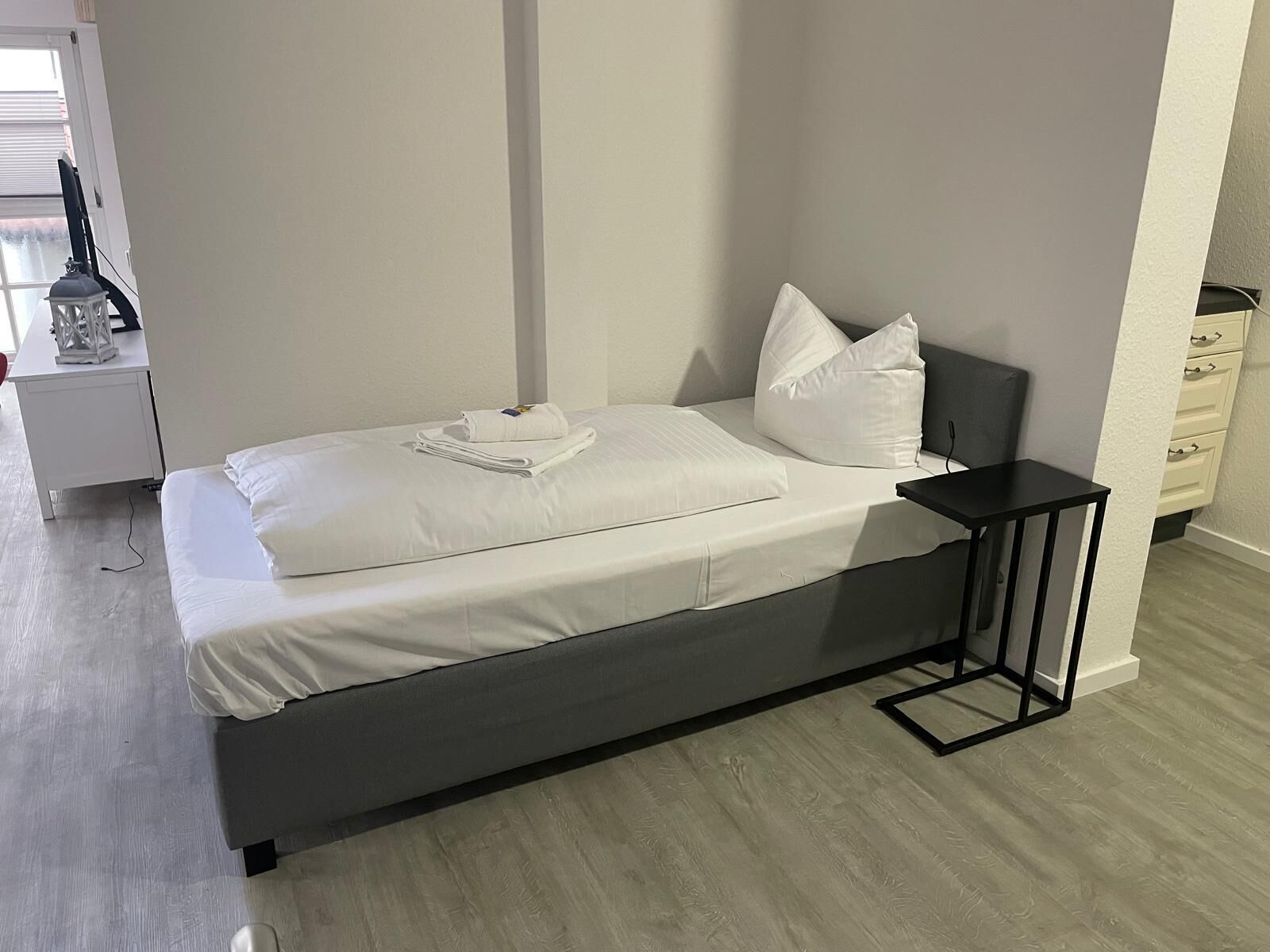Una cama bien hecha con sábanas y almohadas blancas, una mesita de noche con lámpara, y un aparador con cajones.