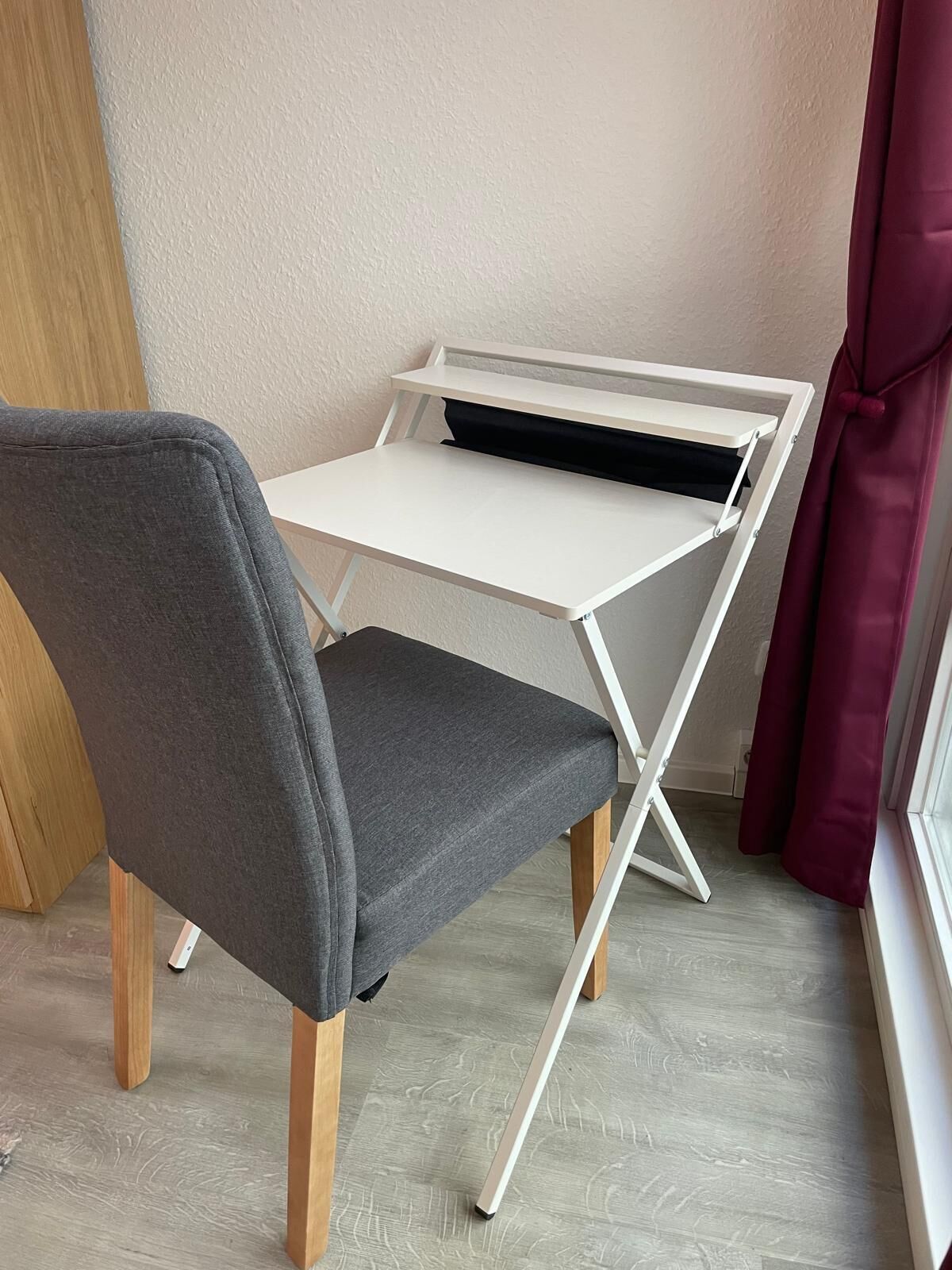 Un escritorio blanco plegable con una silla gris en una habitación con suelo de madera clara y una pared lisa.