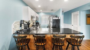 A kitchen with a black countertop, white cabinets, and a stainless steel refrigerator. There are four wooden chairs around the counter.