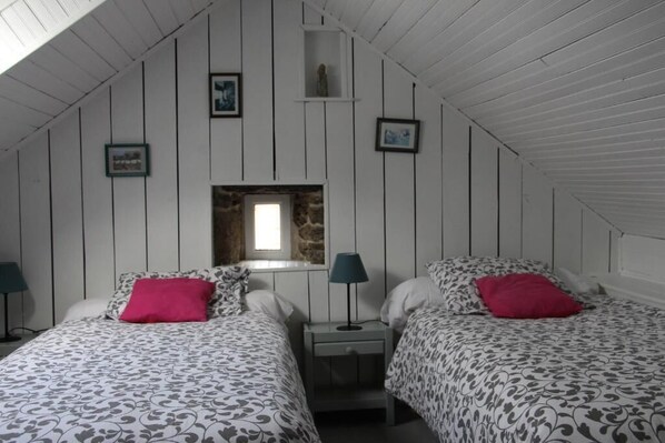 Una habitación con dos camas, cada una con ropa de cama estampada y un cojín rojo. Hay una mesita de noche con una lámpara entre las camas. Las paredes son de color blanco y tienen paneles, y el techo es inclinado.