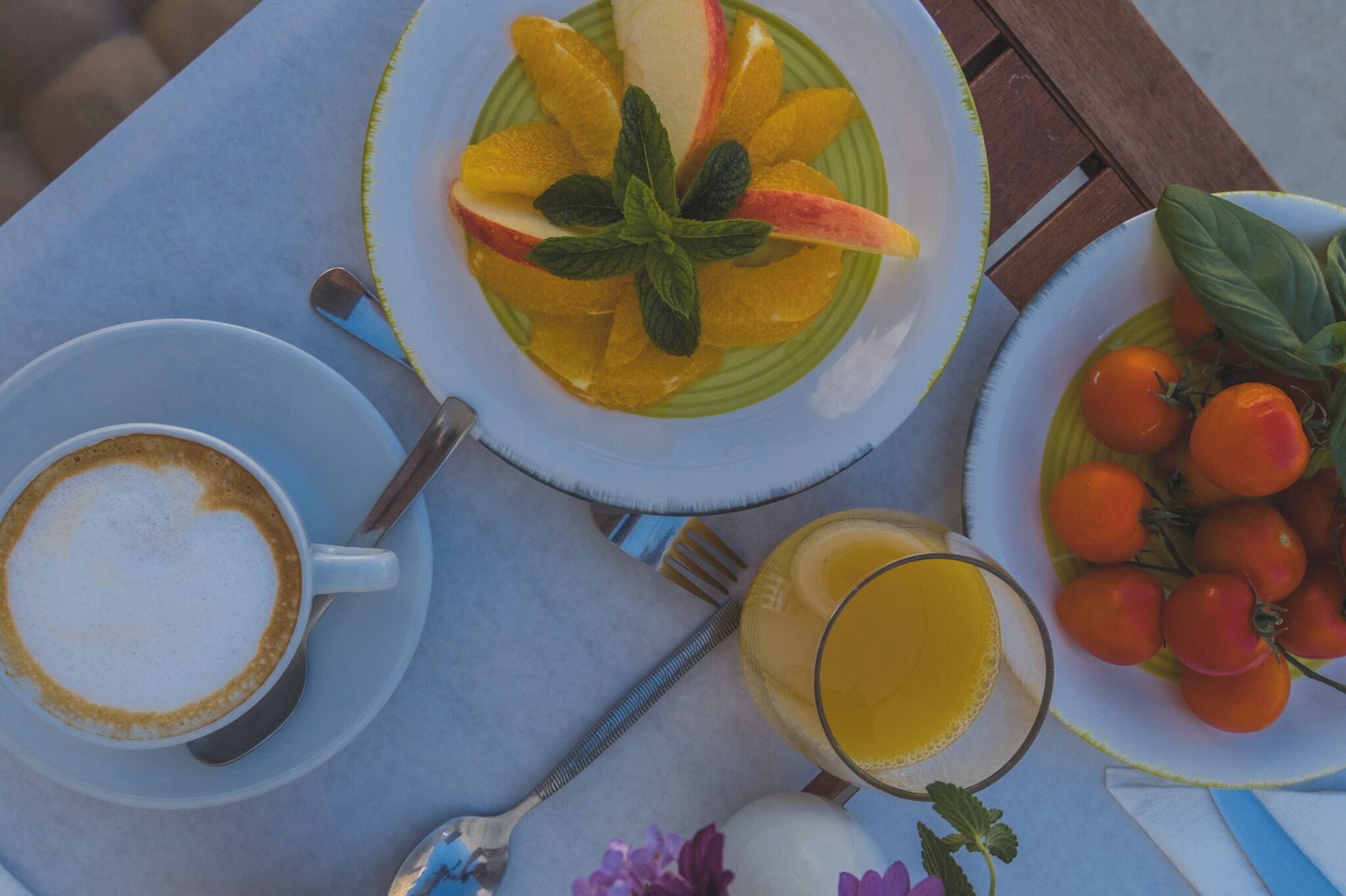 Une table avec une assiette de fruits coupés, un verre de jus d’orange, une tasse de café et une assiette de tomates.