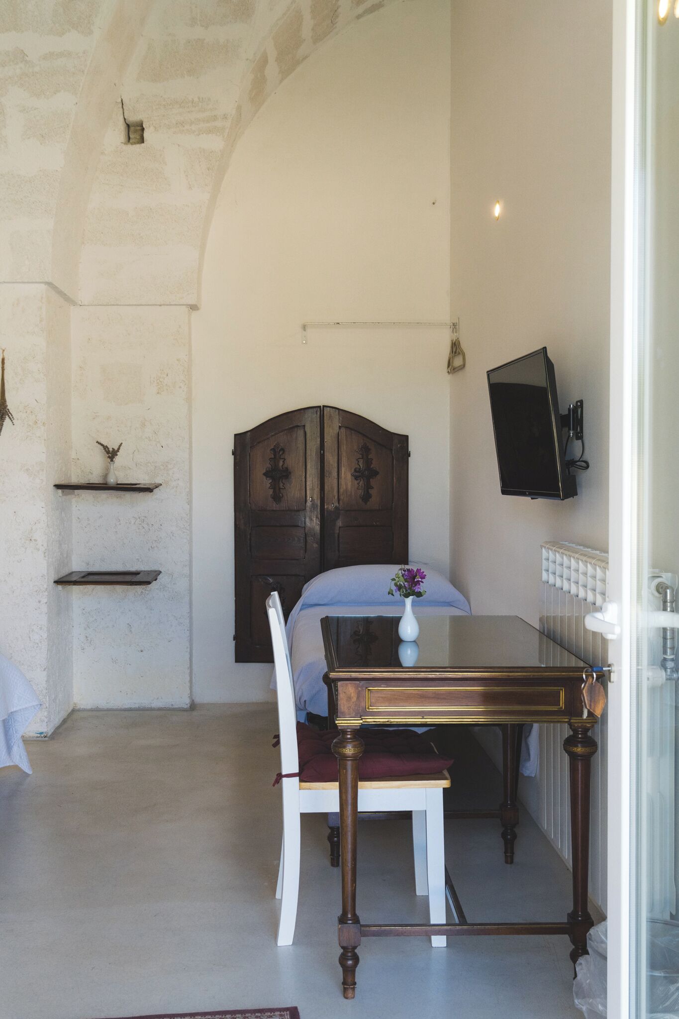 Una habitación con una cama, una mesa de madera con un florero, una silla, un televisor montado en la pared y un radiador.