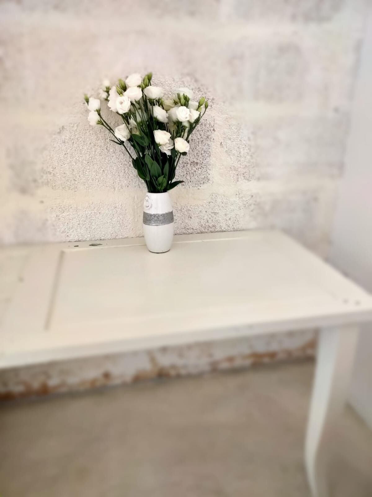 Una mesa blanca con un jarrón de flores blancas frente a una pared con textura.