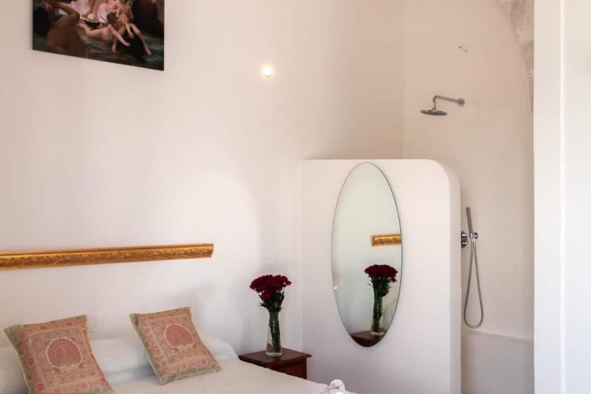 Un dormitorio con cama, banca, espejo, mesita de noche y un cuadro en la pared.