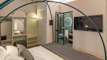 A bedroom with a bed, a small seating area, a walk-in closet, a bathroom with a glass door, and a TV mounted on the wall.