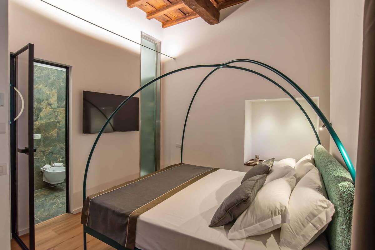 A modern bedroom with a glass-walled shower, a bed with a curved headboard, and a mounted TV.