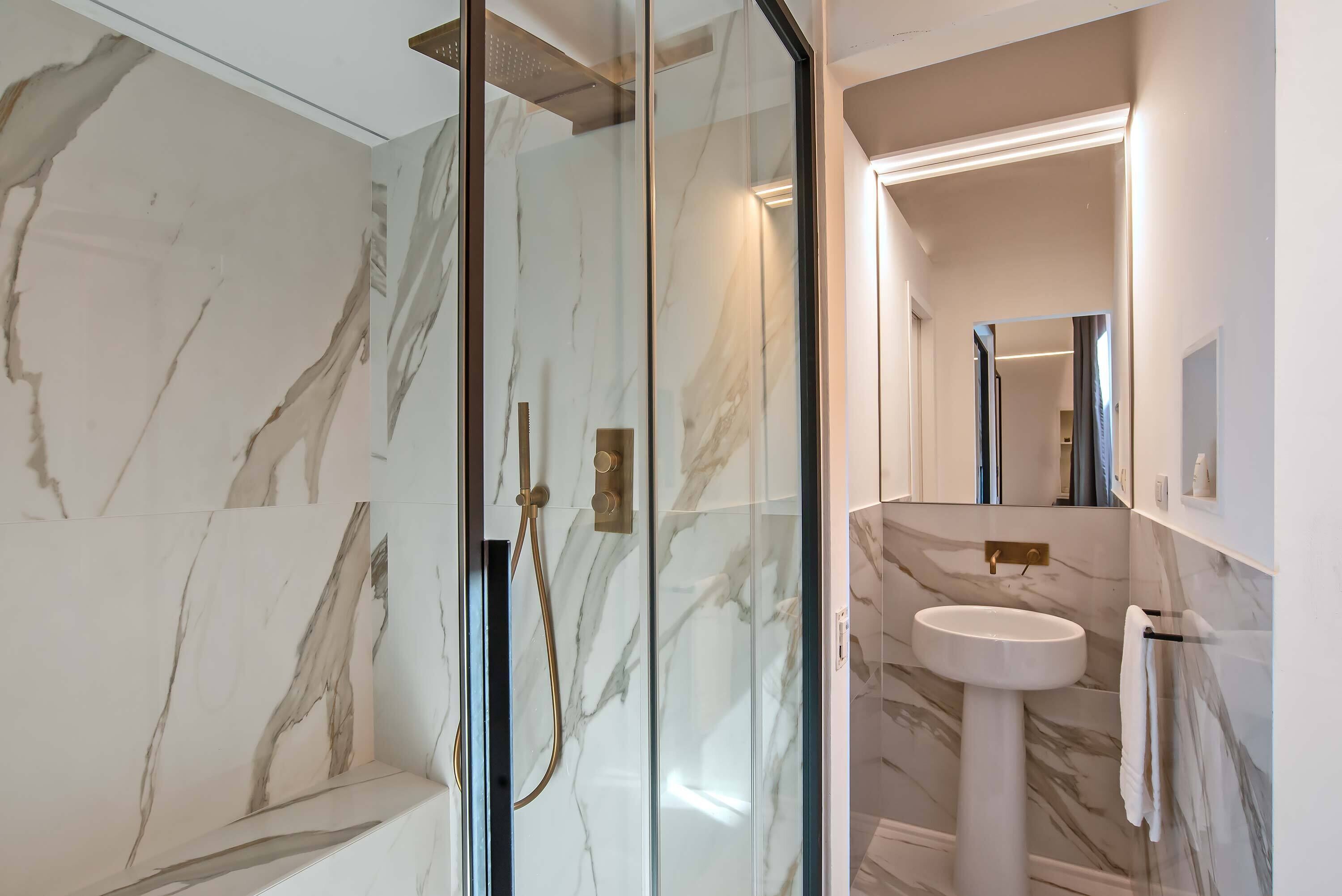 A modern bathroom with a glass-enclosed shower, a marble-tiled wall, a pedestal sink, and a mirror above the sink.