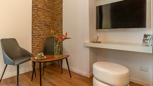 A modern living room with a stone accent wall, a flat-screen TV mounted above a white shelf, and two chairs around a small wooden table with a vase of flowers. - Ora Acanfora Suites (Rome)