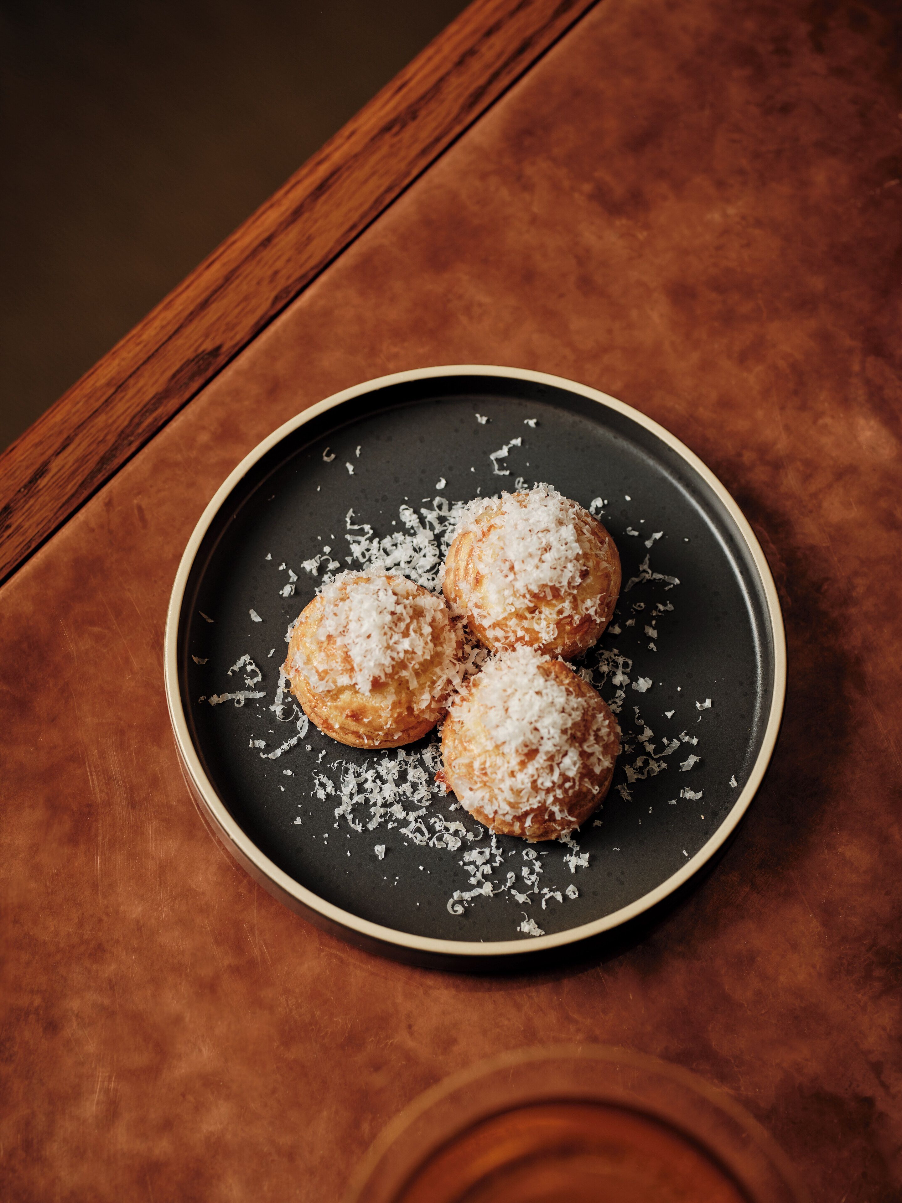 A plate of three round pastries with shredded coconut on a dark plate, placed on a wooden table.