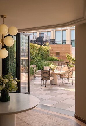 A modern outdoor dining area with a round table and chairs, surrounded by lush greenery and a view of the cityscape. - The Newman (London)