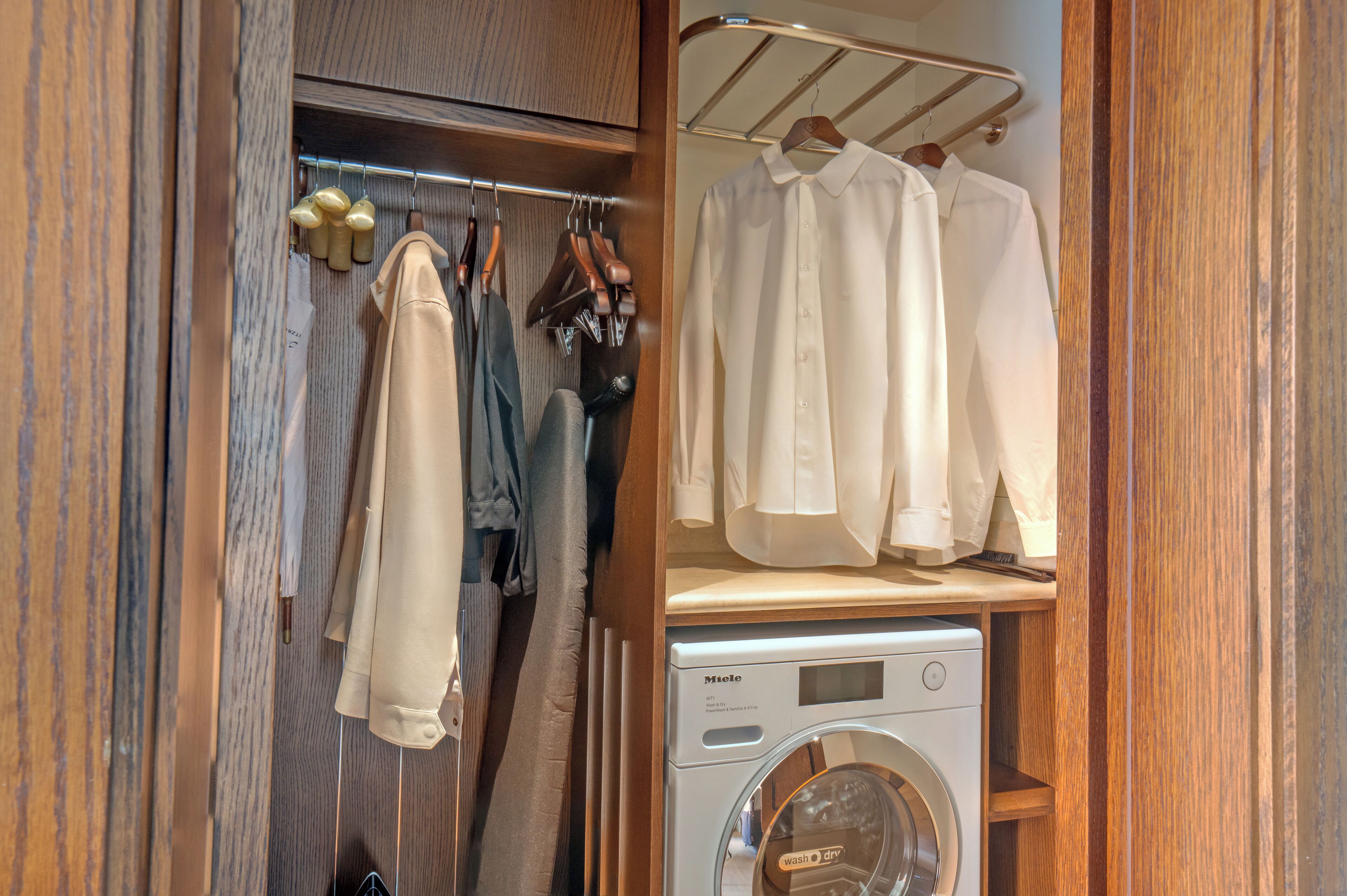A compact closet with hanging clothes, a wooden shelf with folded shirts, and a front-loading washing machine.