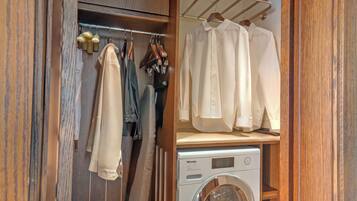 A compact closet with hanging clothes, a wooden shelf with folded shirts, and a front-loading washing machine.