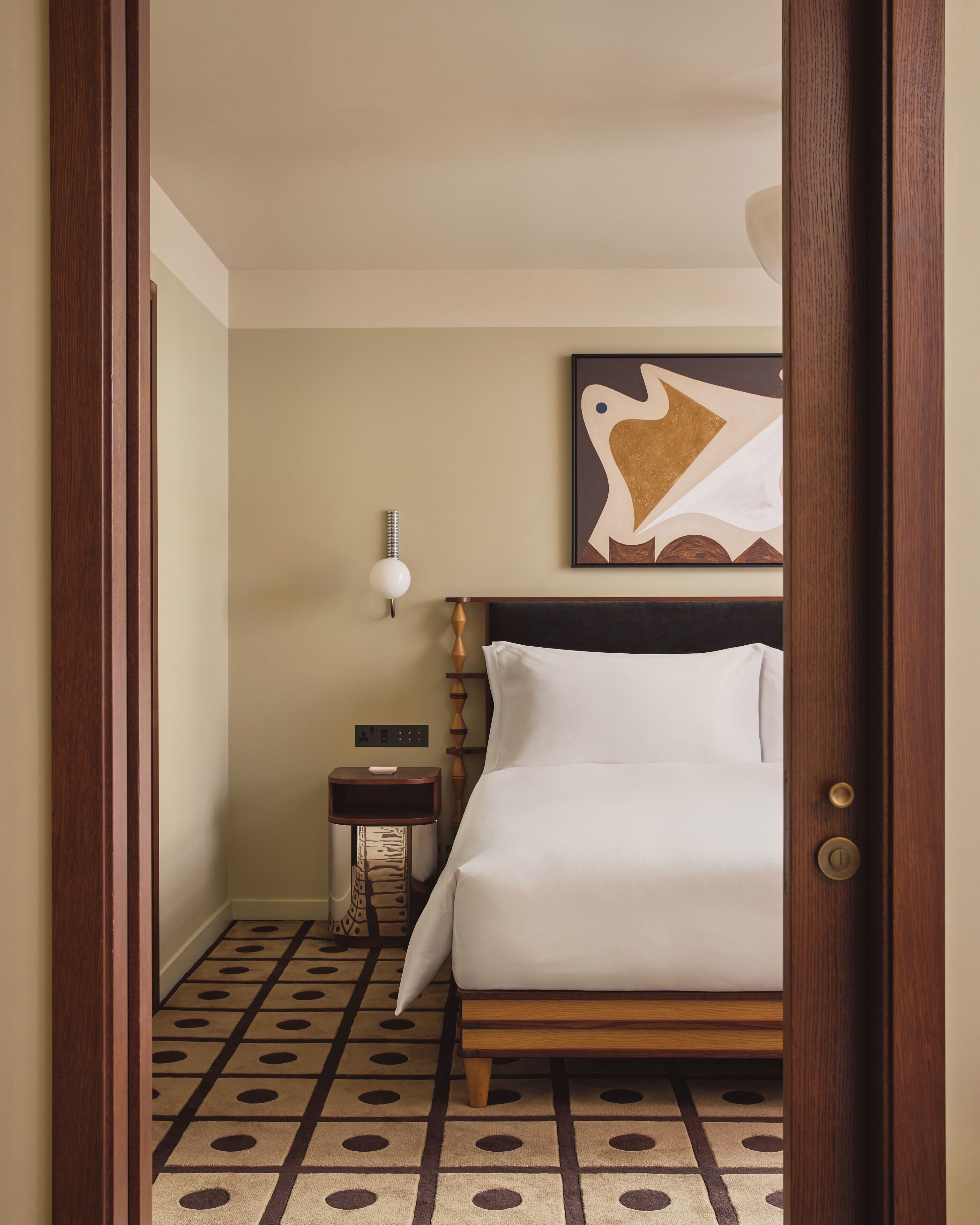 A hotel room with a bed, a nightstand, a wall-mounted lamp, and a painting.