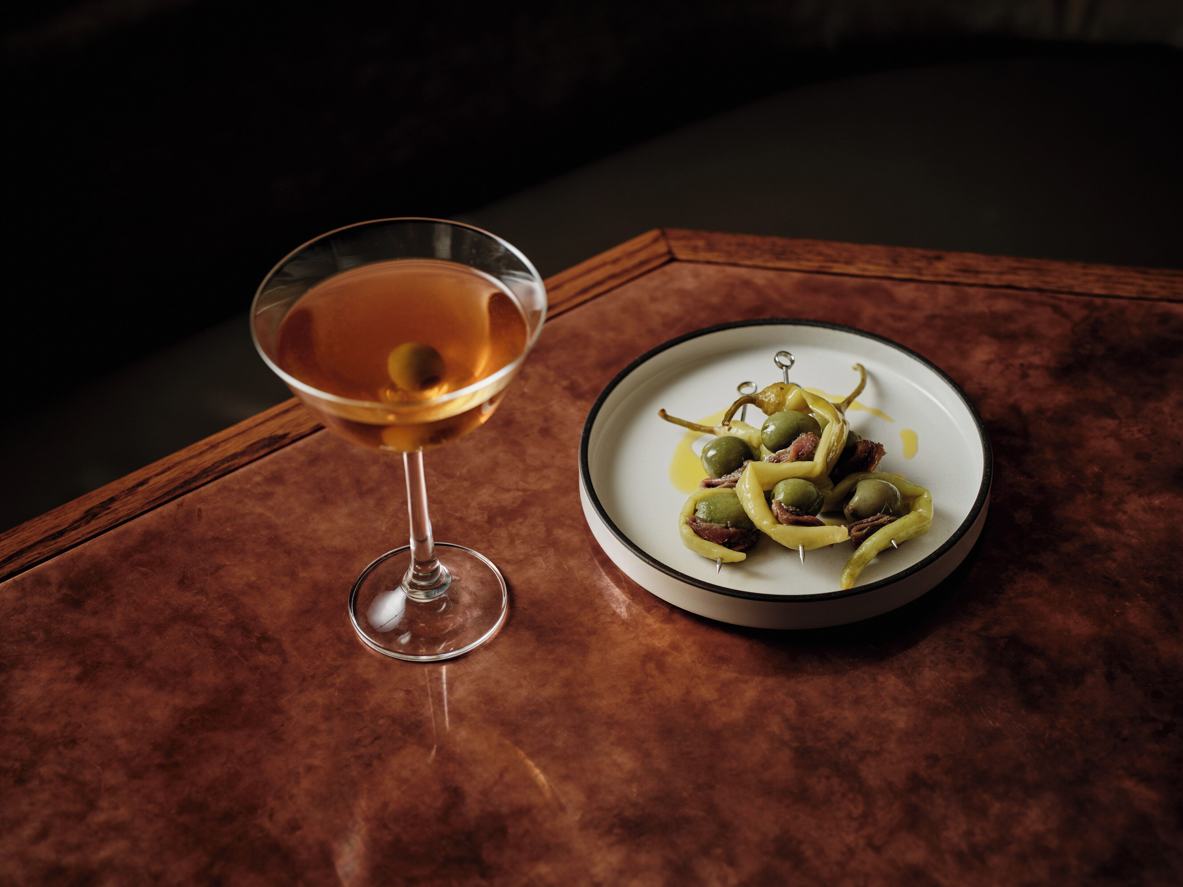 A martini glass with a clear liquid and a plate of olives with a yellow sauce.