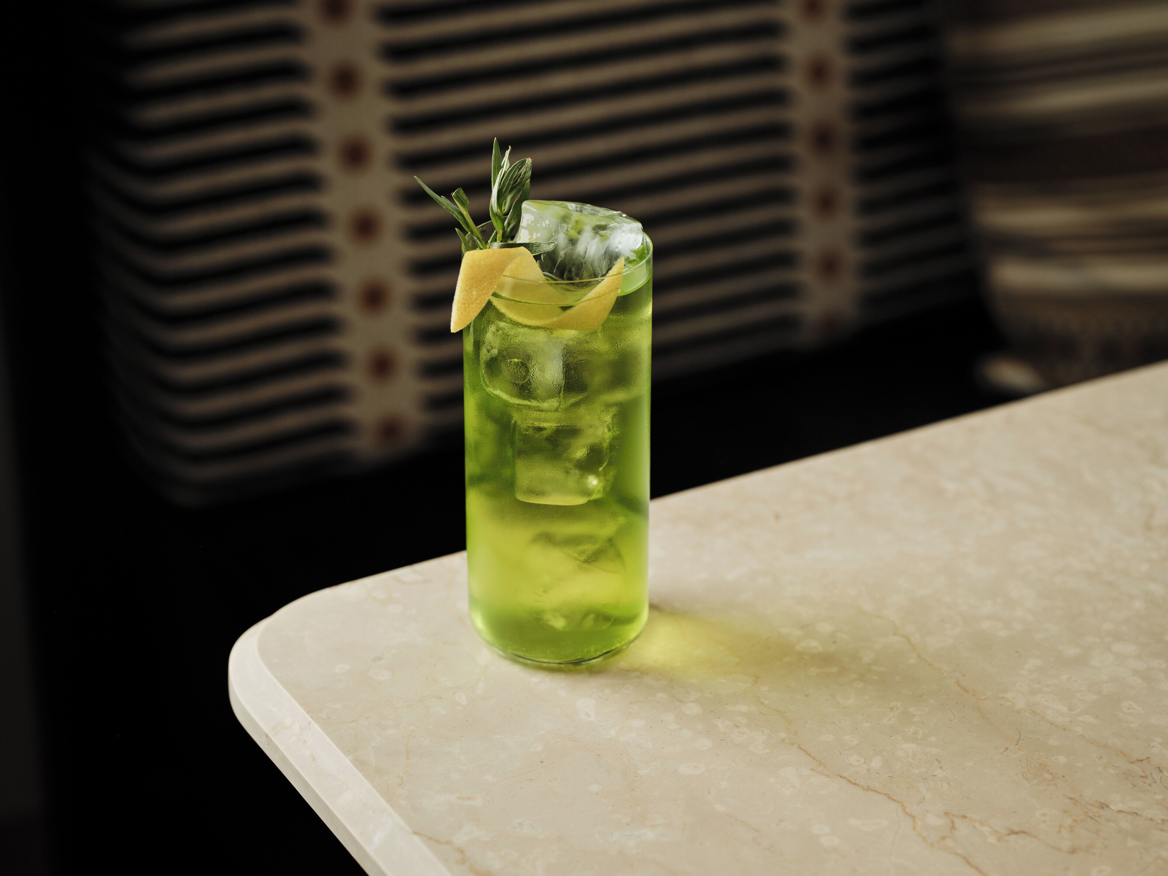 A glass of green beverage with a lemon slice and a sprig of rosemary on a marble countertop.