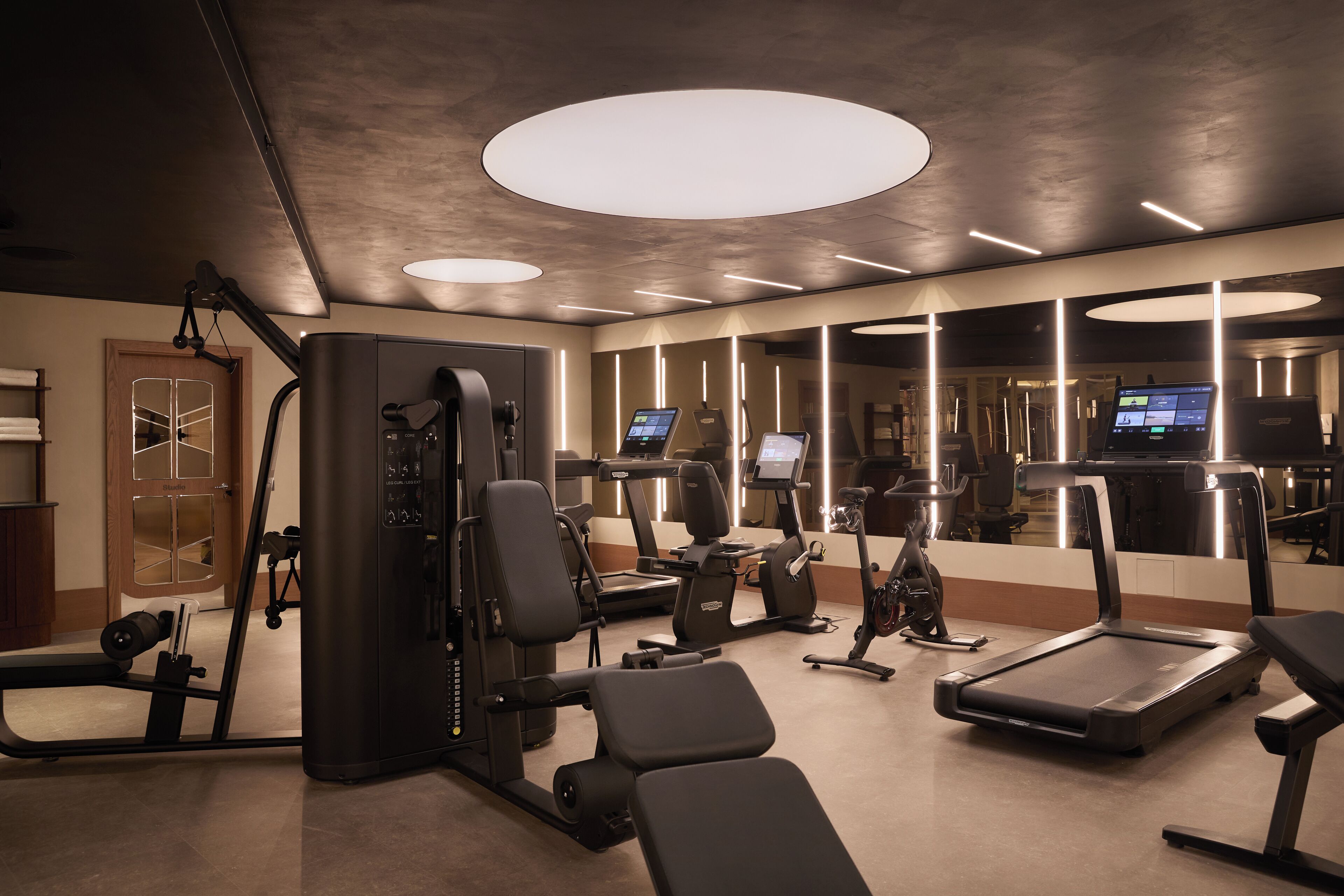 Un gimnasio con diverso equipo de ejercicio, incluyendo cintas de correr y máquinas de pesas, y un espejo grande que refleja el espacio.