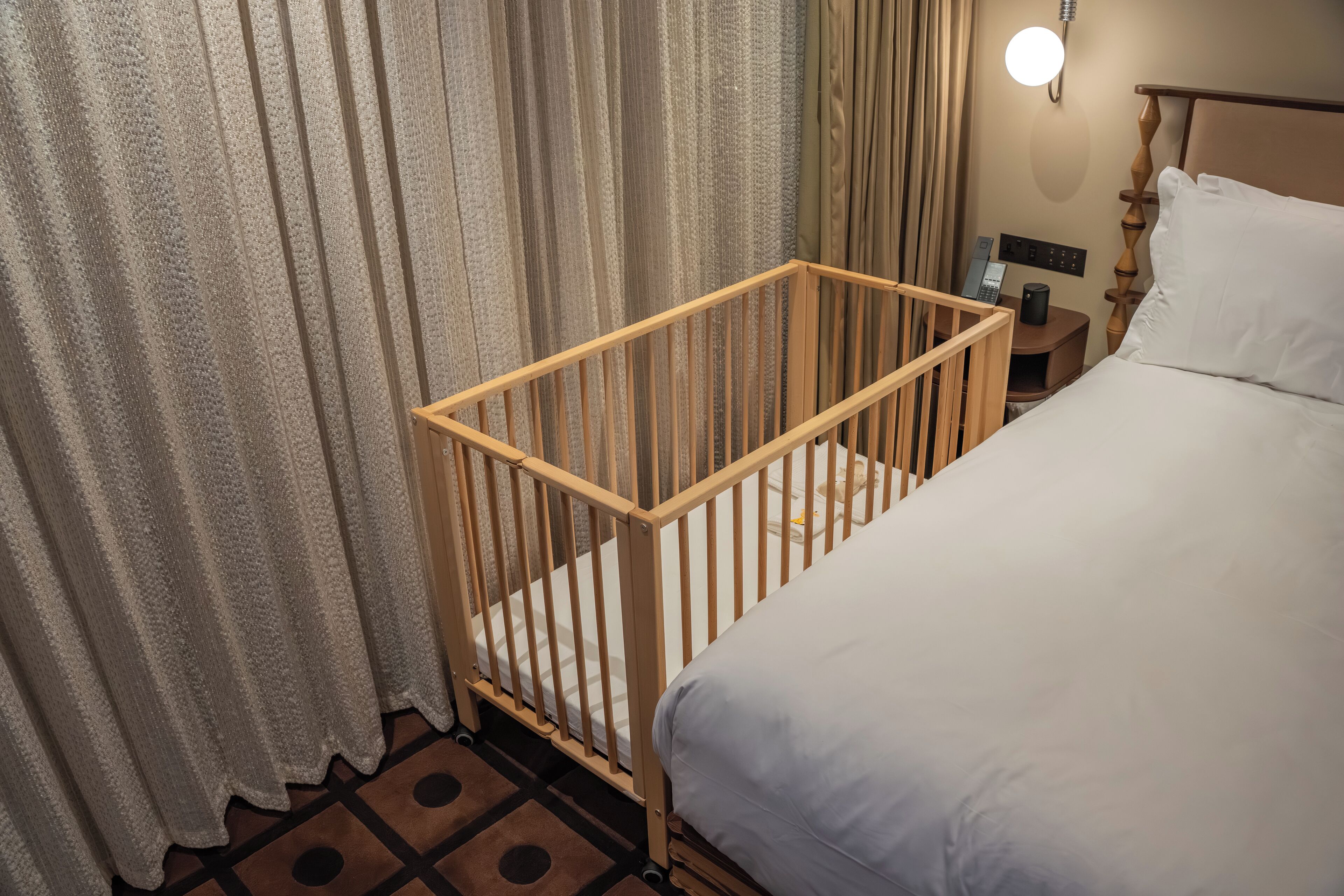 A room with a wooden crib, a bed with white bedding, a nightstand with a lamp, and a curtain.