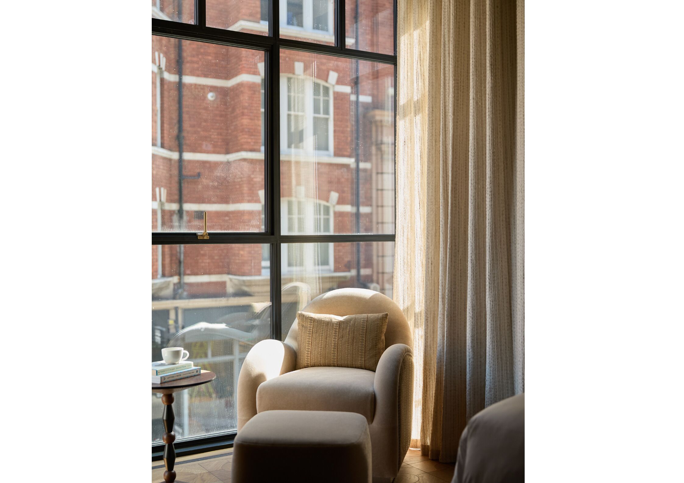 A room with a large window, a chair, a small table with a cup and books, and a curtain.