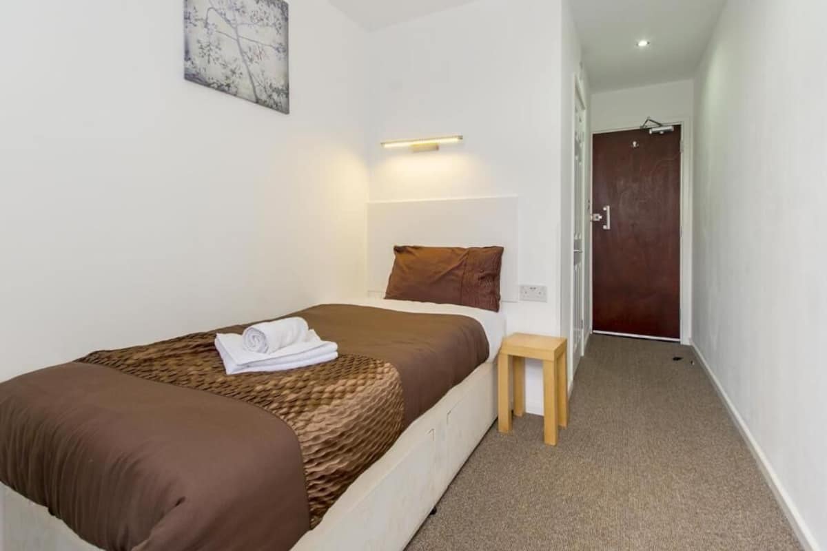 A neatly made bed with a brown blanket and pillows, a small wooden stool, a wall-mounted light, and a door in the background.