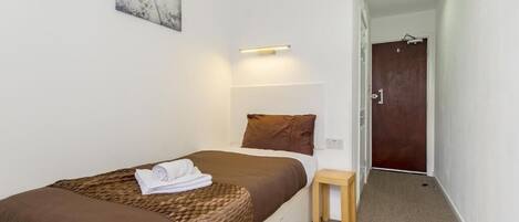 A neatly made bed with a brown blanket and pillows, a small wooden stool, a wall-mounted light, and a door in the background.