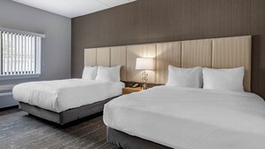 Une chambre d’hôtel avec deux lits, une tête de lit en bois, une table de chevet avec une lampe et une fenêtre avec des stores.