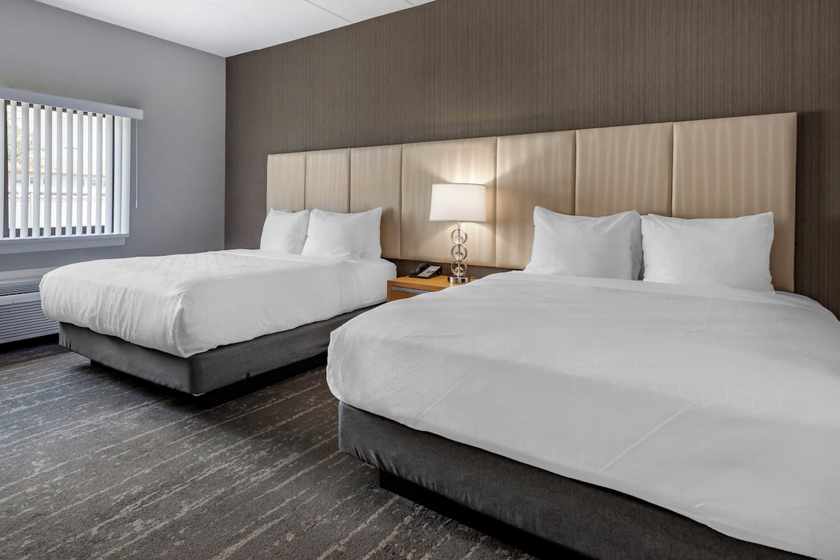Une chambre d’hôtel avec deux lits, une tête de lit en bois, une table de chevet avec une lampe et une fenêtre avec des stores.