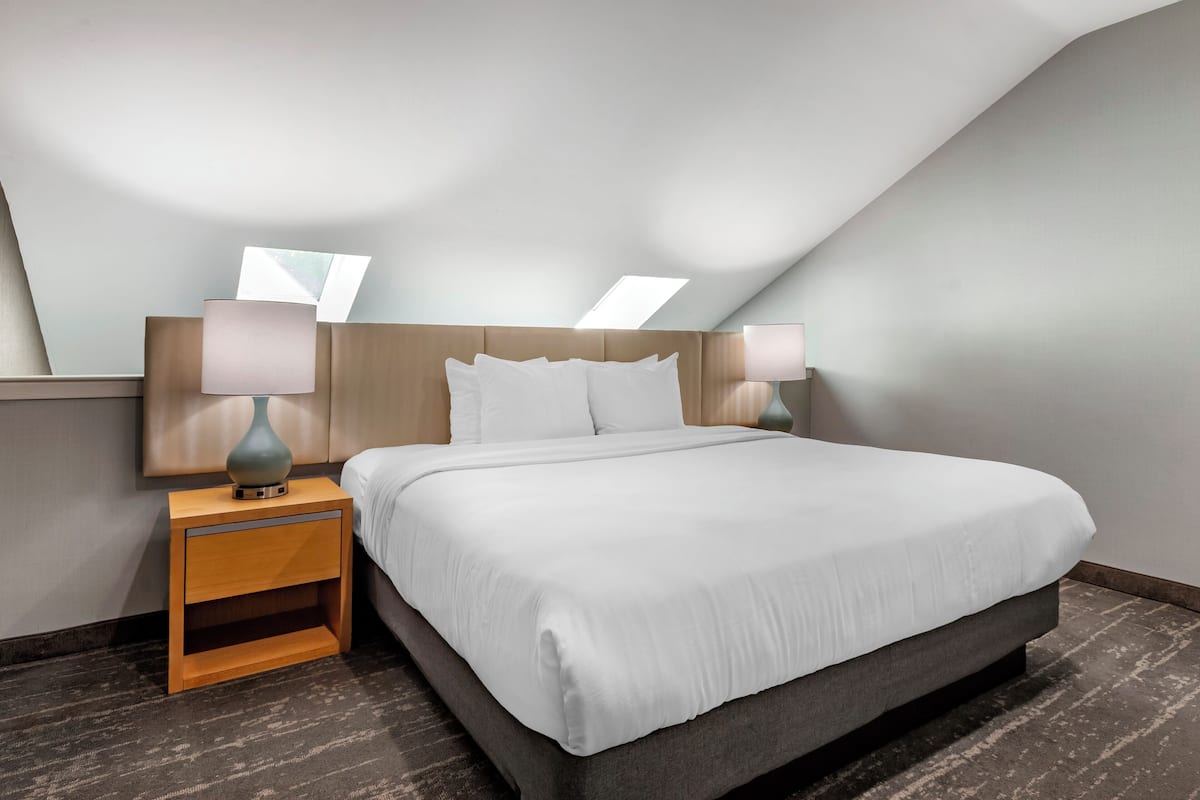 Un lit bien fait dans une chambre d’hôtel, avec une table de chevet en bois, deux lampes de chevet et un plafond en pente.