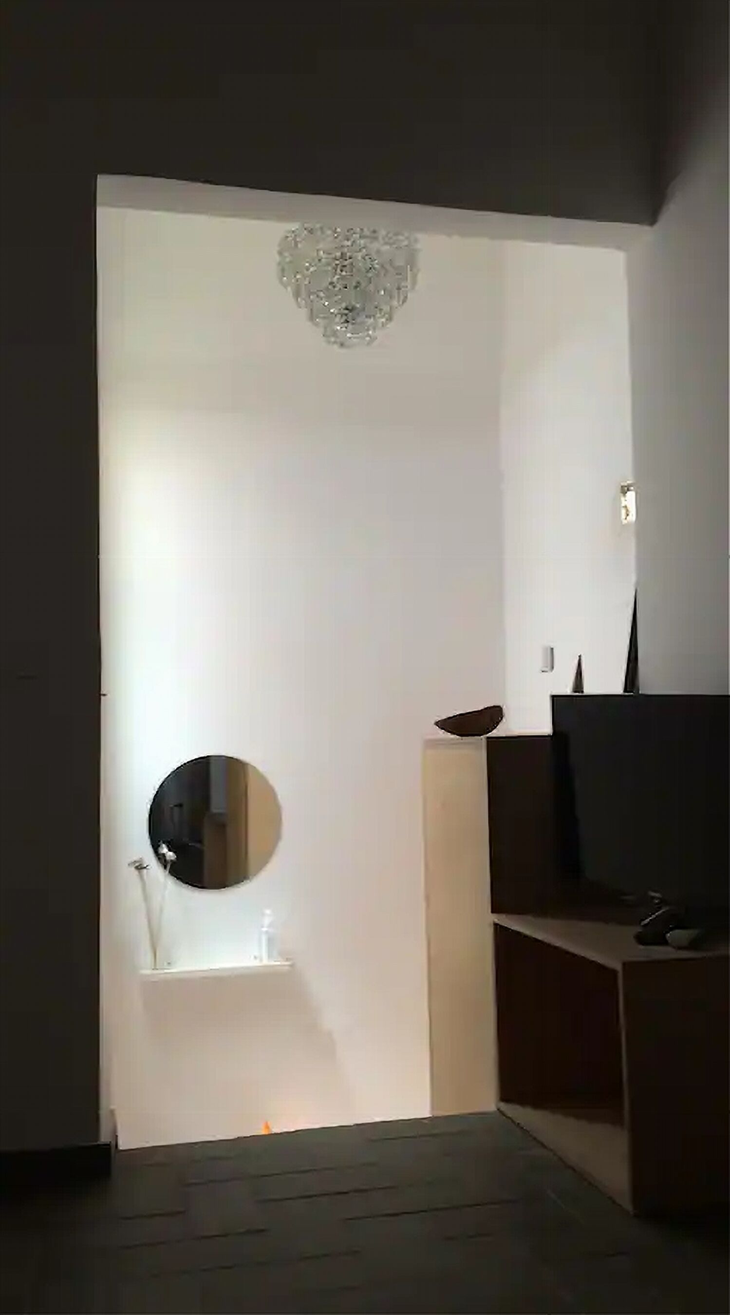 Una habitación con una puerta blanca, un espejo redondo, un lavamanos pequeño y un televisor sobre un soporte de madera.