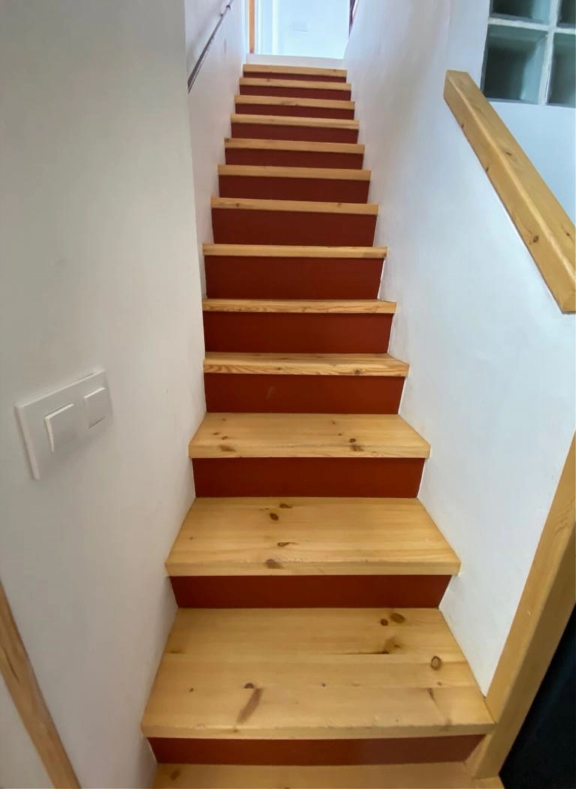 Een houten trap met een leuning aan de rechterkant en een lichtschakelaar aan de linkerkant.