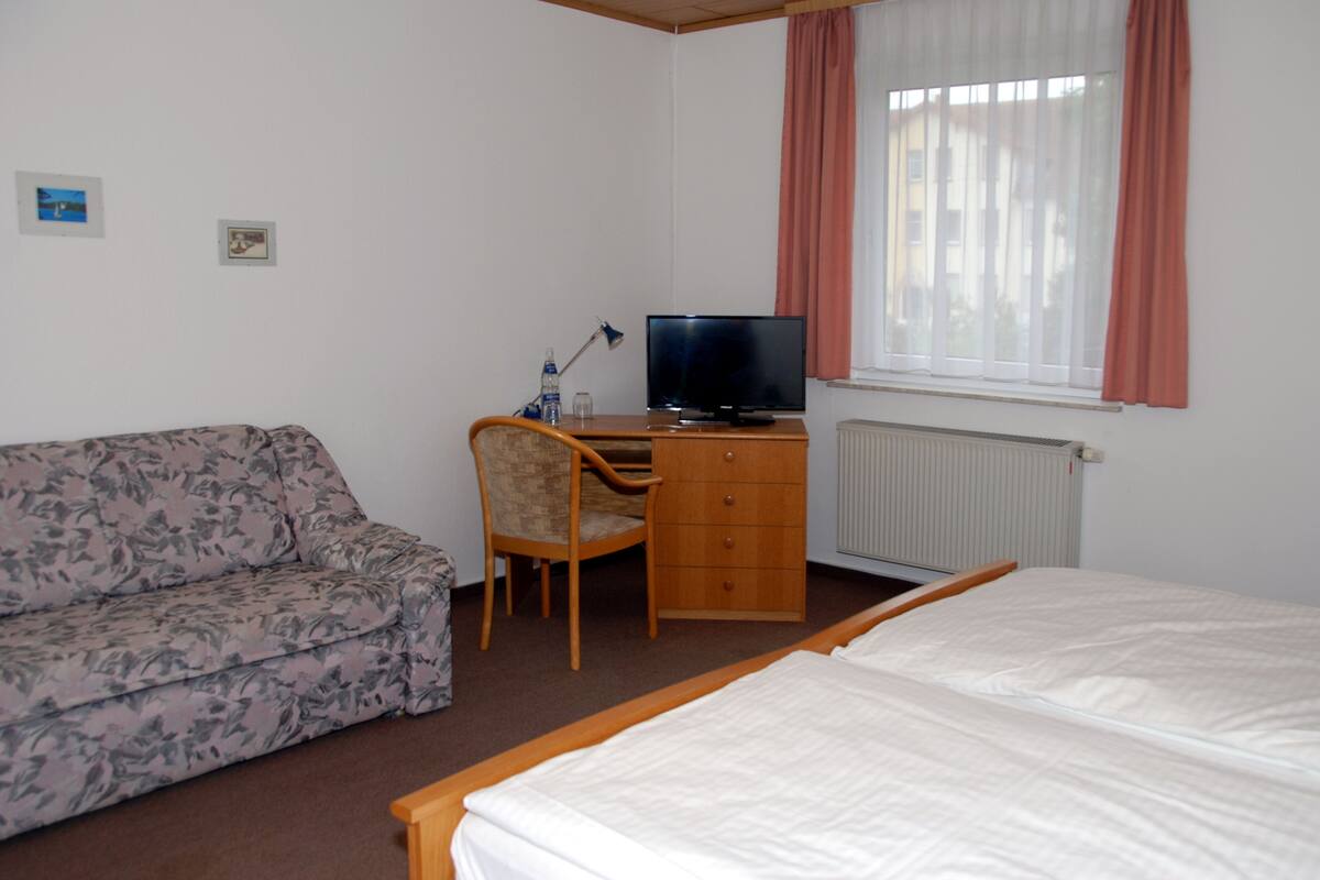 A hotel room with a bed, a desk with a computer, a chair, a sofa, and a window with curtains.