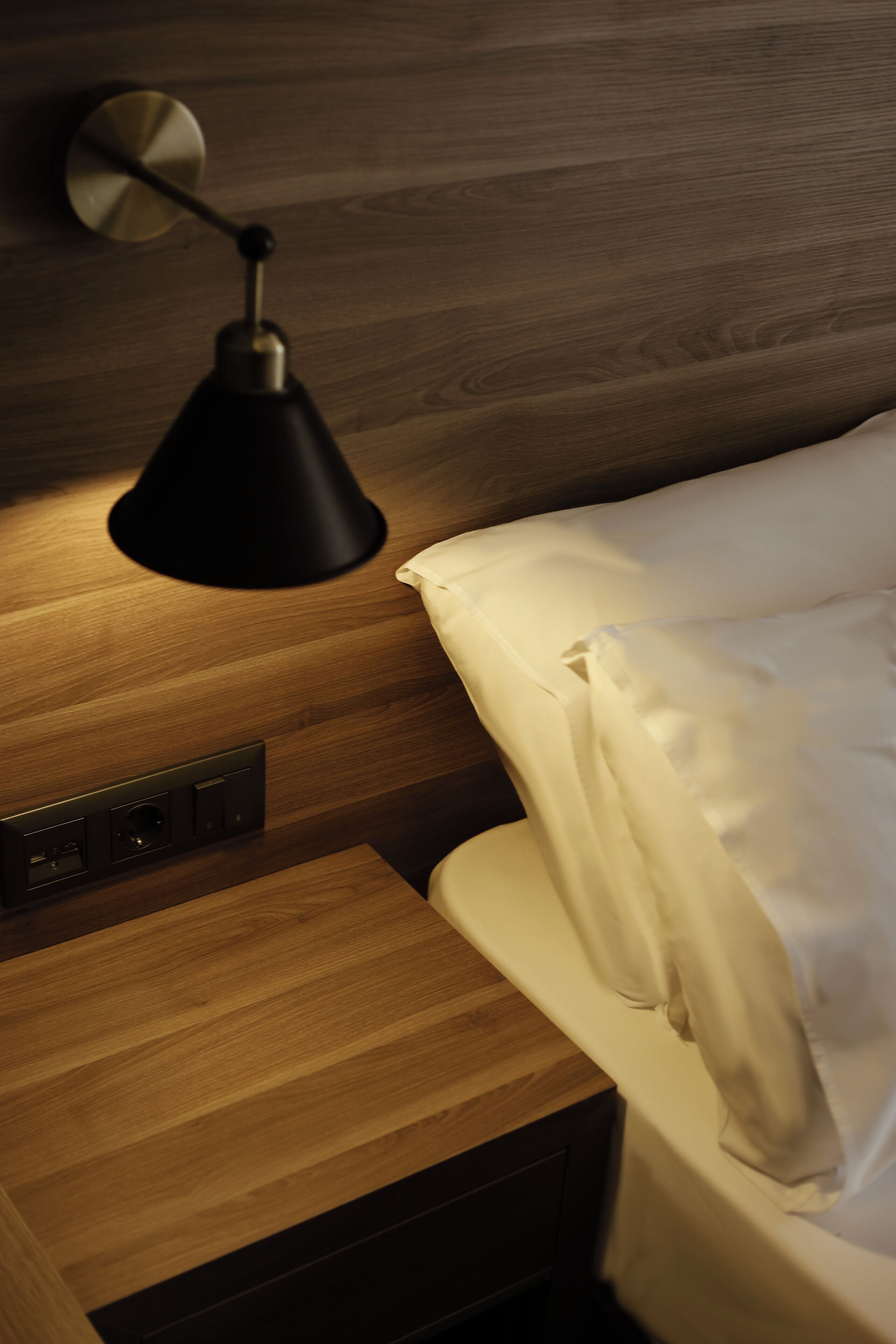A wooden bedside table with a black lamp, a power outlet, and a white bed with a white pillow.