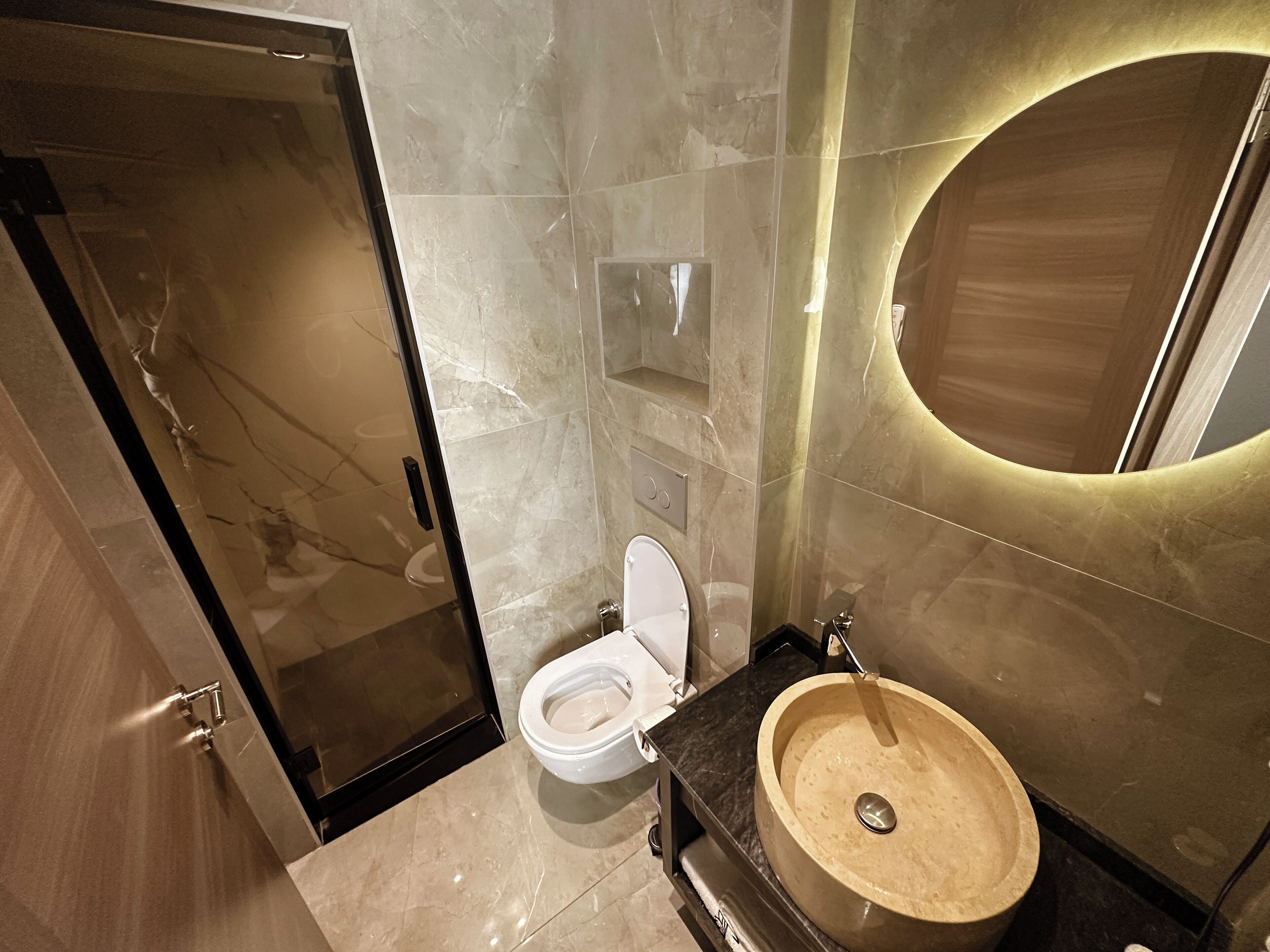 Une salle de bain moderne avec un sol en marbre, des toilettes, un lavabo avec un récipient en bois et un grand miroir doté d’un cadre circulaire.