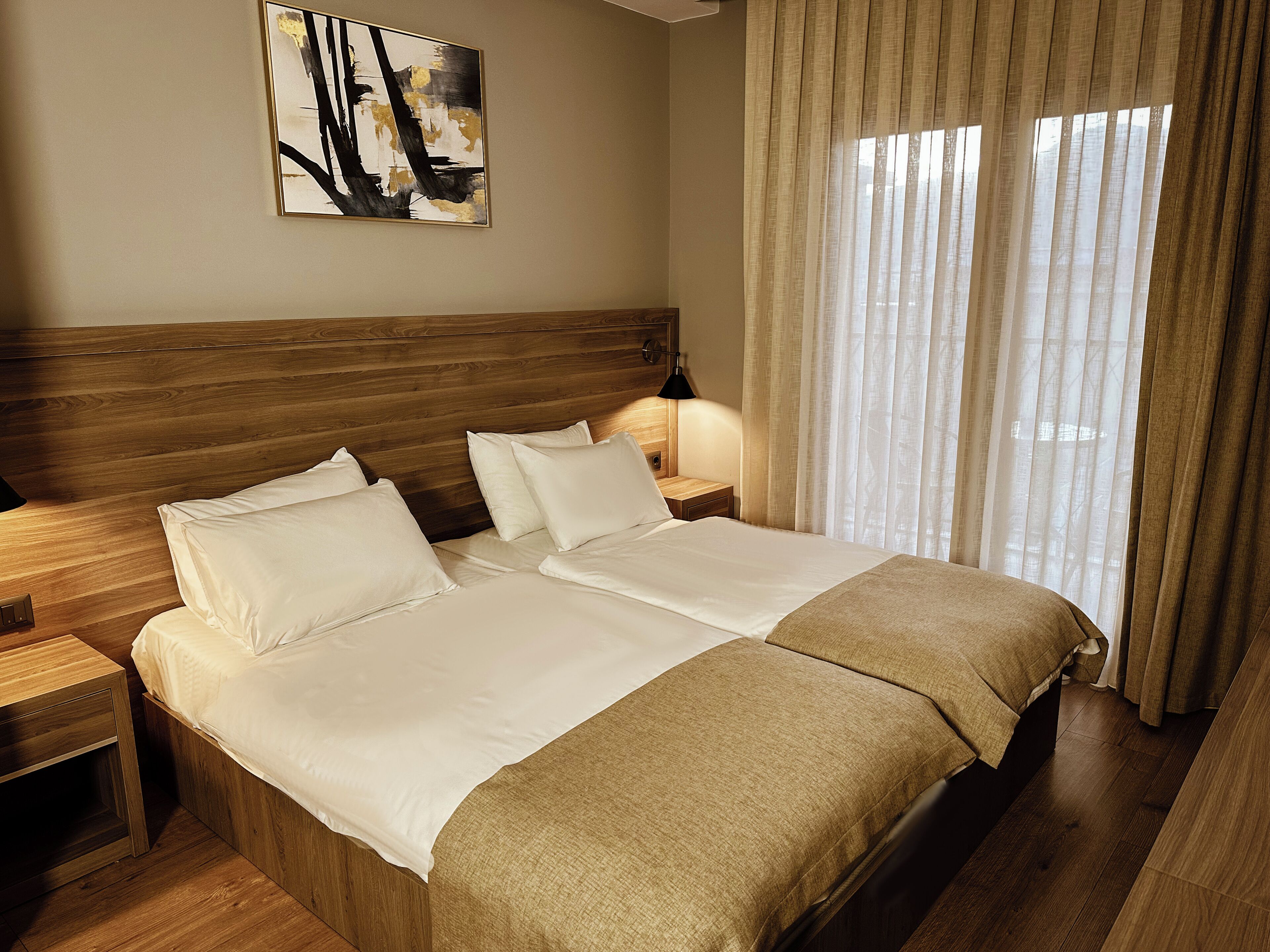 A double bed with a wooden headboard, two bedside tables, a lamp, and a large window with sheer curtains.