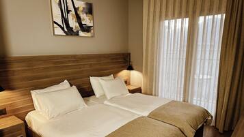 A double bed with a wooden headboard, two bedside tables, a lamp, and a large window with sheer curtains.