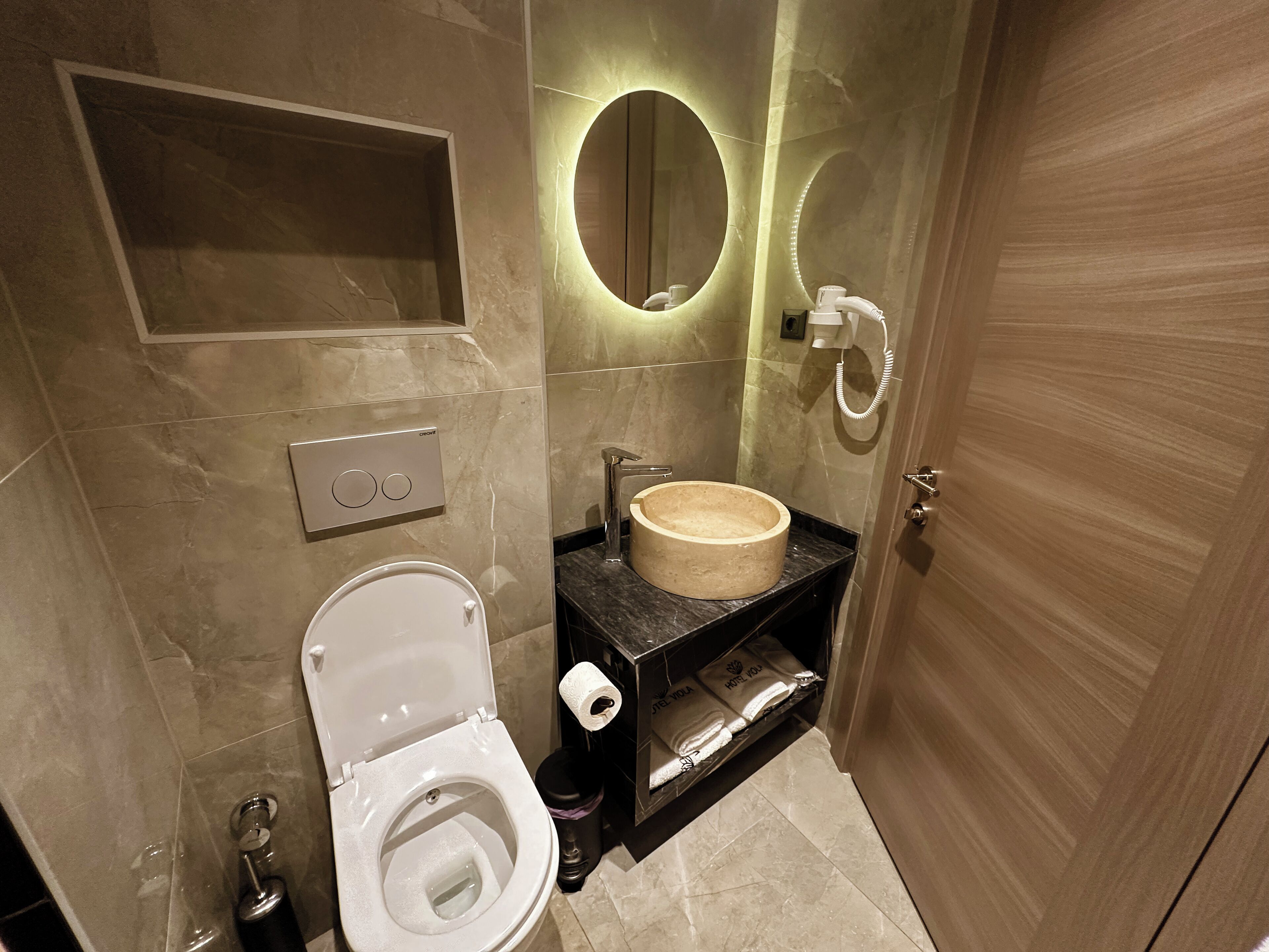 Une salle de bain moderne comprenant des toilettes, un lavabo avec un receveur en marbre, un miroir avec un éclairage circulaire et un porte-serviettes.