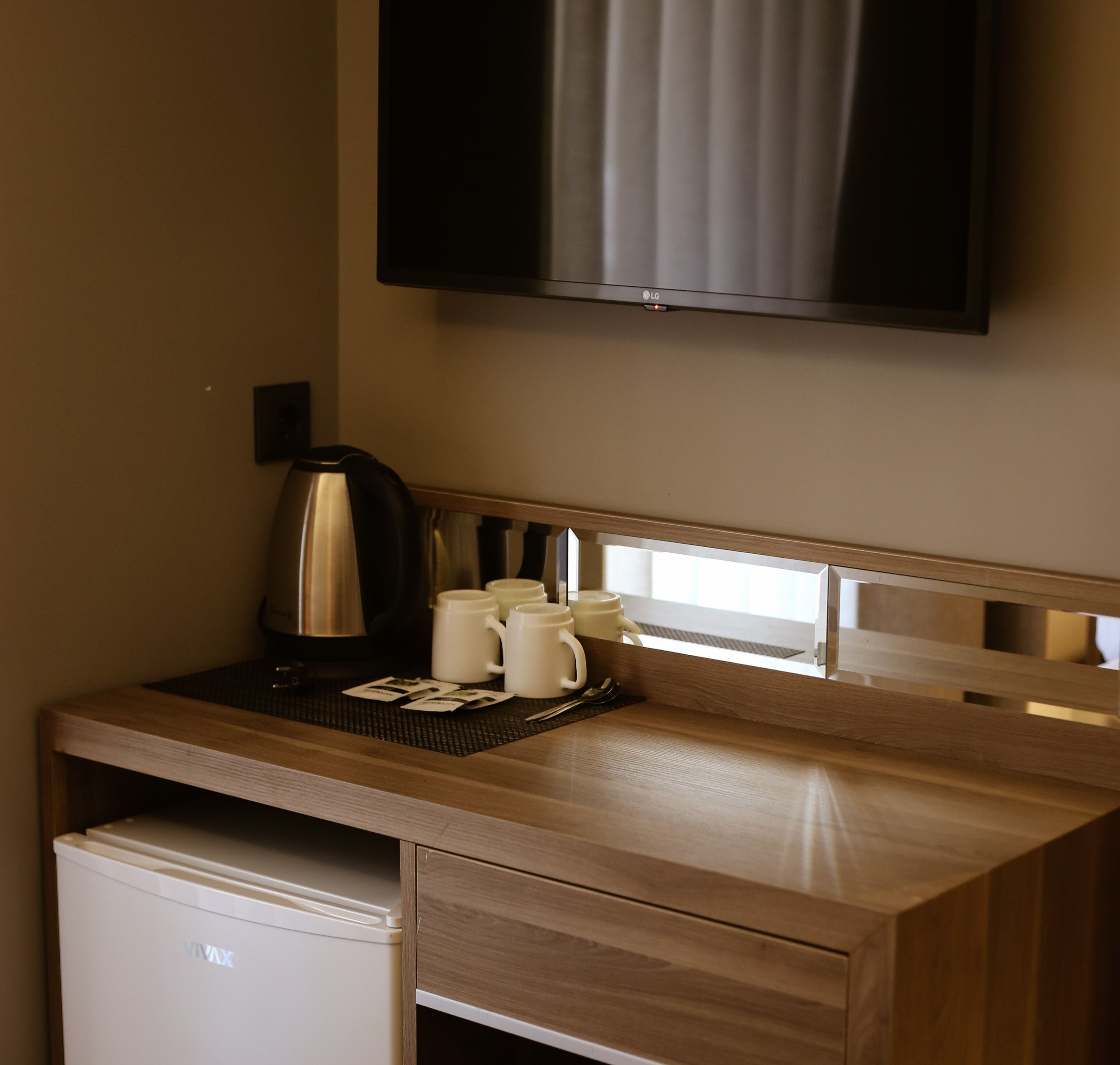 A hotel room with a wooden desk, a coffee maker, four white mugs, a small refrigerator, and a flat-screen TV mounted on the wall.
