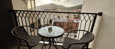 A balcony with two wicker chairs and a small table, overlooking a cityscape with buildings and a mountain.