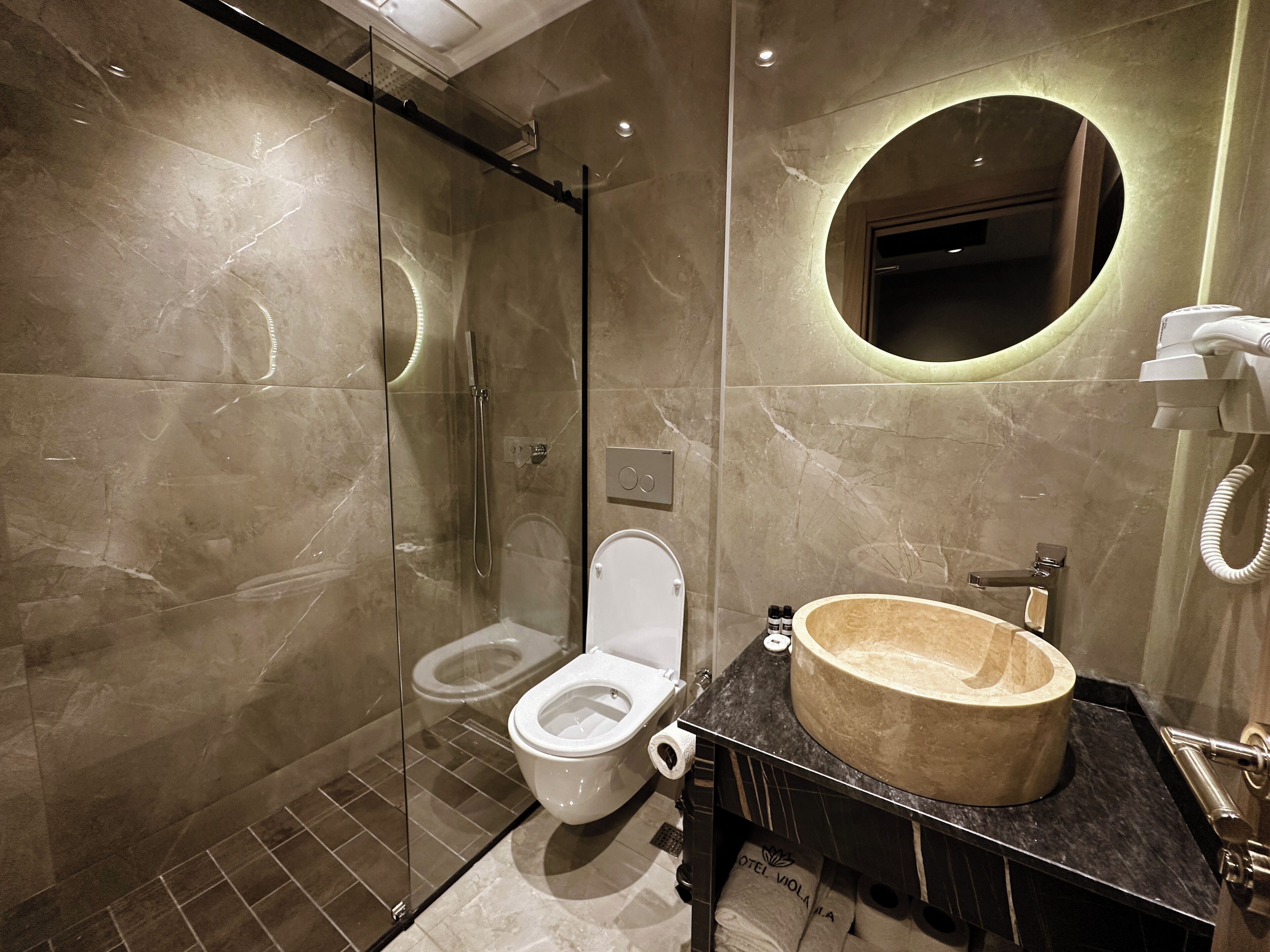 Un baño moderno con ducha envuelta en vidrio, un lavamanos redondo con una pila de mármol y un inodoro con tapa blanca.